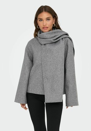 Manteau en mélange de laine gris avec un col drapé et surdimensionné, des manches larges, une fermeture avant asymétrique et une texture douce. Associé à un pantalon noir.