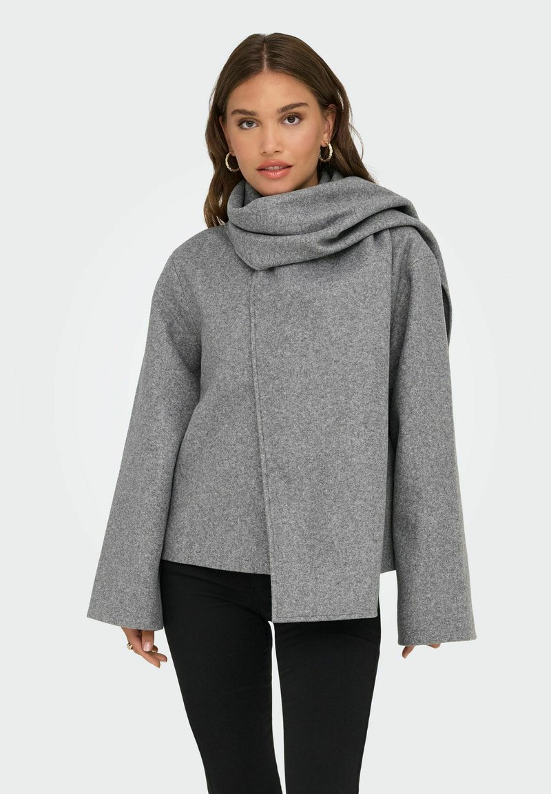 Grå ullblandad kappa med ett draperat, överdimensionerat krage, vida ärmar, asymmetrisk framknäppning och mjuk textur. Stylerad med svarta byxor.