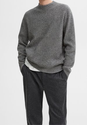 Homme portant un pull en maille côtelée gris par-dessus une chemise blanche, avec un pantalon gris foncé à fines rayures, mains dans les poches, sur fond uni.