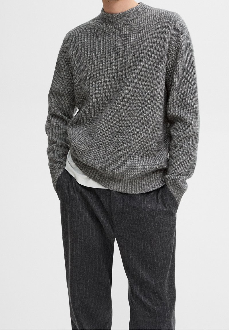 Homme portant un pull en maille côtelée gris par-dessus une chemise blanche, avec un pantalon gris foncé à fines rayures, mains dans les poches, sur fond uni.