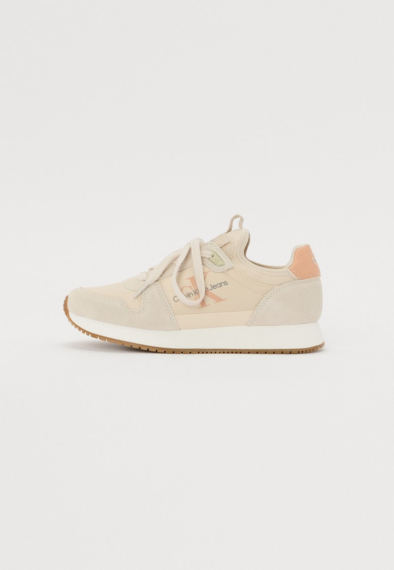 Beige athletic sneaker made of suede and mesh, featuring a rounded toe, lace-up front, and contrasting light orange accents on heel.