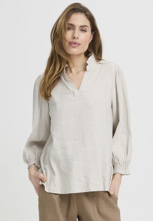 Žena s dlhými vlasmi, oblečená v svetlobéžovej blúzke s volánikovým výstrihom do V a rukávmi, v kombinácii s voľnými hnedými nohavicami, stojaca s rukami vreckách.