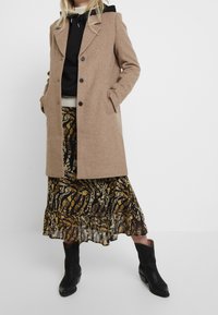 Donna che indossa un cappotto beige con bottoni, felpa nera con cappuccio, gonna midi a fantasia e stivali neri alla caviglia, in piedi con le mani nelle tasche.