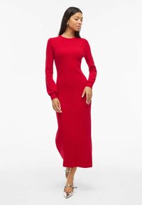 Abito midi rosso lavorato a maglia con maniche lunghe, corpetto aderente, scollatura rotonda e texture liscia, abbinato a décolleté metallici con punta affusolata.