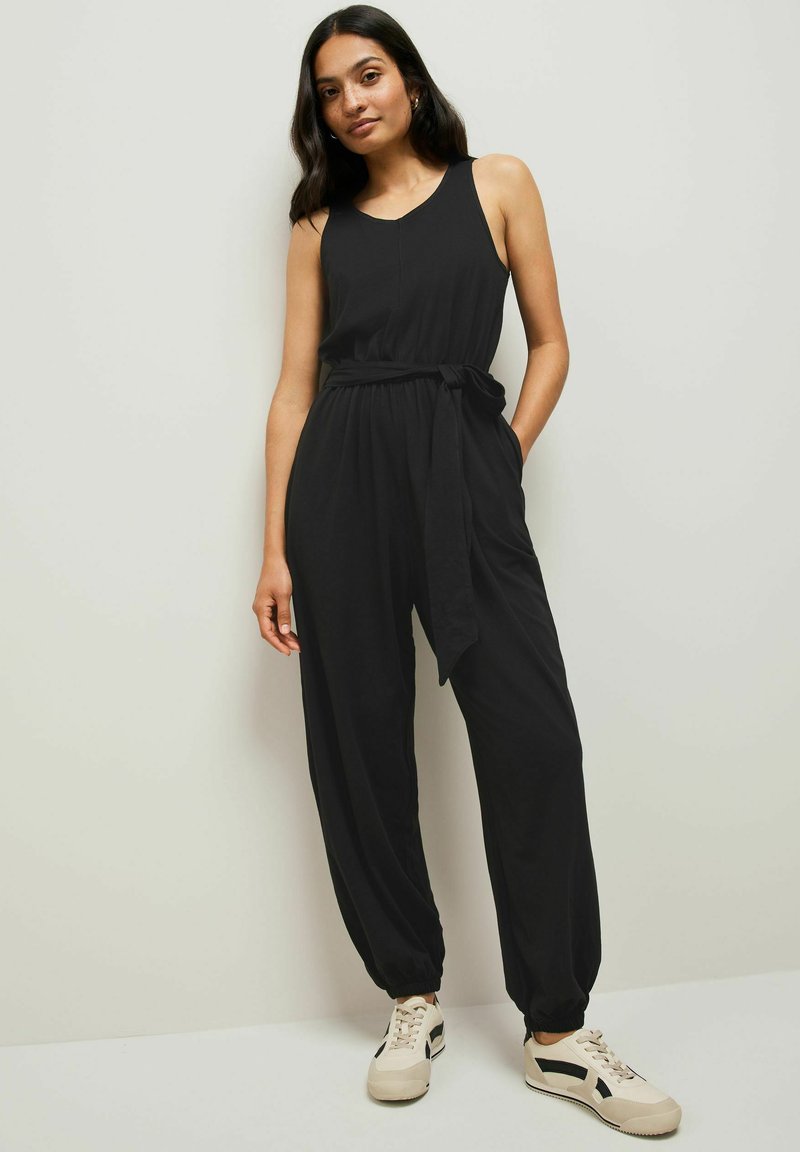 Zwarte mouwloze jumpsuit met een relaxte pasvorm, een strikceintuur in de taille en taps toelopende broekspijpen. Gepaarde met beige sneakers met zwarte accenten.