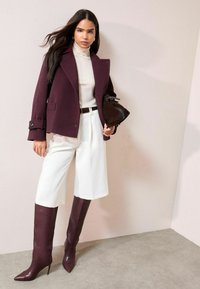 Bordo spalvos dvieilis vilnos mišinys švarkas, kreminės spalvos golfas, balti plisuoti kuločių tipo šortai, bordo spalvos aulinukai iki kelių su smailiu priekis ir juoda rankinė.