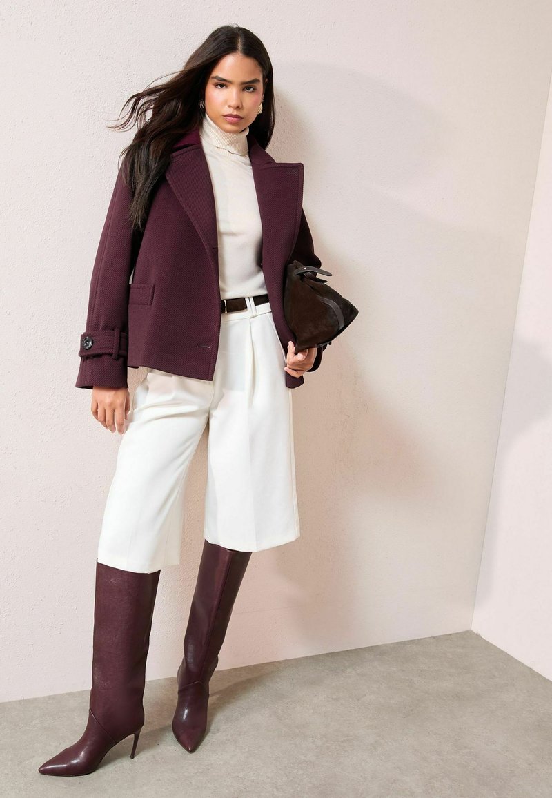 Bordo spalvos dvieilis vilnos mišinys švarkas, kreminės spalvos golfas, balti plisuoti kuločių tipo šortai, bordo spalvos aulinukai iki kelių su smailiu priekis ir juoda rankinė.