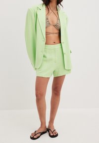 Hellgrüner maßgeschneiderter Blazer mit oversized Passform, passende Shorts und ein beiger Bikini-Top mit dünnen Trägern, getragen mit schwarzen Flip-Flops.