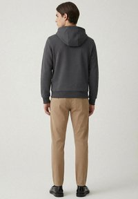 Mörkgrå luvtröja med kenguruficka, kombinerad med ljusbruna slim fit-byxor och svarta skor, sedd bakifrån.