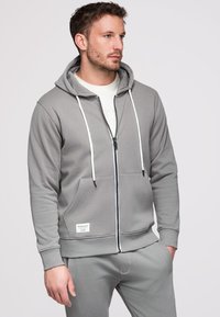 Homme portant un sweat à capuche gris zippé avec des cordons blancs et un pantalon de jogging gris assorti sur un fond clair uni.