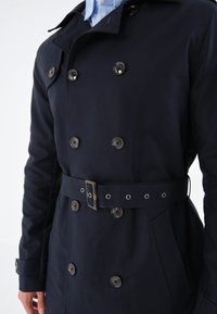 Navyblå trenchcoat gjord av slätt tyg, med dubbelknäppta knappar, midjebälte och sidofickor.