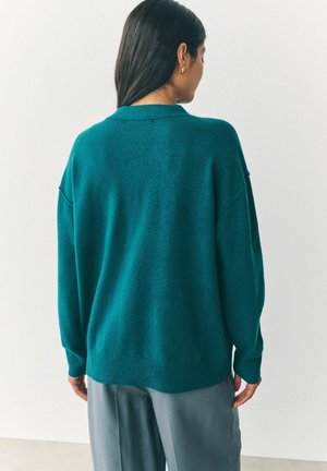 Tyrkysový oversized svetr s límcem, dlouhými rukávy a texturovaným pletením. Částečně zasunutý do světle šedých kalhot. Jednoduchý design a hladký povrch.