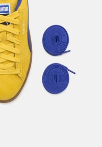 Yellow suede sneaker with blue accents and brown rubber sole, paired with two rolled blue shoelaces.