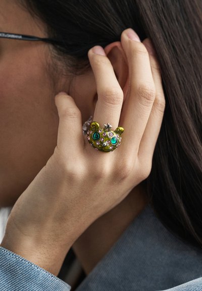 Green animal-shaped ring with blue and pink stones, featuring a star accent. Worn on the index finger, background shows part of a denim sleeve.