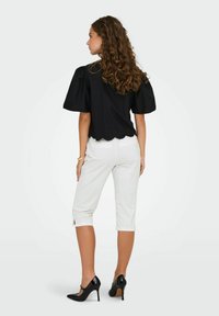 Crna bluzica s napuhanim rukavima i valovitim rubom, u kombinaciji s bijelim skraćenim hlačama s prorezima na manžetama i crnim cipelama na visoku petu.