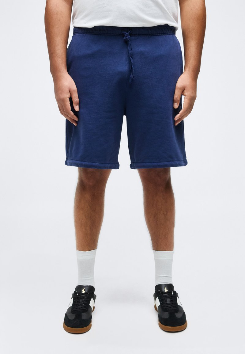 Άντρας που φοράει ναυτικό μπλε σόρτς με κορδόνι, λευκές κάλτσες και μαύρα αθλητικά παπούτσια, στέκεται με τα χέρια στα πλάγια πάνω σε απλό λευκό φόντο.