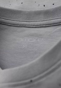 Detalle de una camiseta de algodón gris que muestra un escote redondeado, una textura suave y una etiqueta impresa que dice "Ombre M 148" con pequeñas manchas.