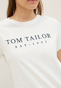 Witte katoenen T-shirt met "TOM TAILOR" en "EST-1962" in marineblauw gedrukt. Ronde halslijn, korte mouwen, gladde textuur.