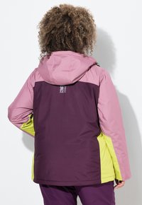 Person mit lockigem Haar trägt eine farblich abgesetzte Jacke in Pink, Lila und Gelb und steht mit dem Rücken zur Kamera vor einem weißen Hintergrund.