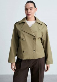 Olive green double-breasted jacket with large lapels, two buttons, and loose sleeves. Worn over a light top, paired with dark trousers.
