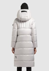 Hosszú, világosszürke, kapucnis pufferkabát, függőleges steppelt mintával és egy logófolttal a szegély közelében. Puha, sima textúra. Fekete kötött sapka.