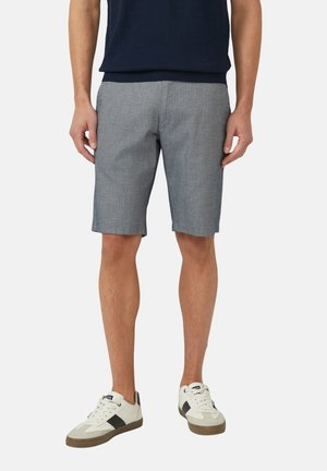 Mann trägt blaue, strukturierte Shorts und weiße Sneakers mit schwarzen Details und steht vor einem einfarbigen Hintergrund.