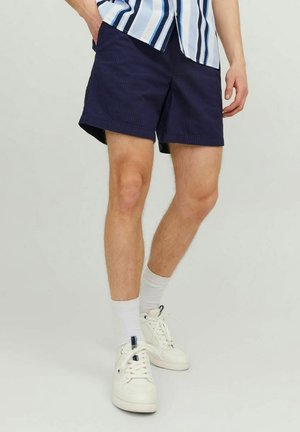 Marineblaue Cordhorts mit lockerem Schnitt, seitlichen Taschen und einem Saum, der über dem Knie endet. Kombiniert mit weißen Turnschuhen und Socken.