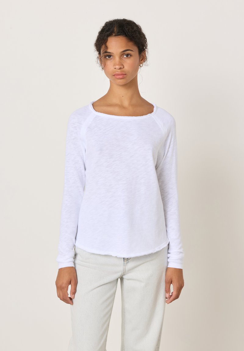 Ung kvinna med lockigt hår, klädd i en vit långärmad tröja och ljusa byxor, stående framför en enkel ljus bakgrund.