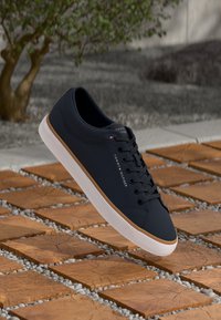 Marineblå Tommy Hilfiger sneakers med hvid sål, svævende over en træflisevej med små grå grusafstande.
