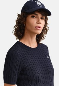 Giovane con capelli ricci indossa un maglione blu navy a maniche corte lavorato a trecce e un berretto da baseball blu navy con testo bianco ricamato.