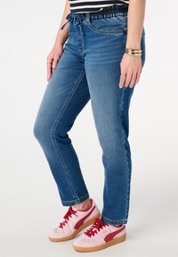 Jean en denim avec un lavage bleu, présentant une taille élastique avec cordon de serrage et une coupe slim. Associés à des baskets roses avec des accents rouges.