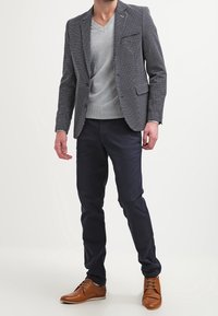 Mężczyzna w szarym swetrze V-neck, wzorzystym żakiecie, ciemnych spodniach i brązowych skórzanych butach eleganckich stoi na tle białym.