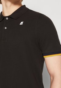 Černé polo tričko s texturovanou tkaninou, které má bílé logo na hrudi a barevné pruhované akcenty na rukávech.