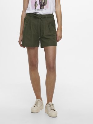 Pantaloni corti verde oliva su misura con pieghe, una cintura e tasche laterali. Abbinati a una t-shirt grafica bianca e sneakers platform beige.