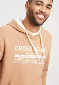 Beżowa bluza z kapturem z białymi sznurkami i nadrukowanym tekstem. Posiada kieszeń kangurkę i luźny krój. Wykonana z miękkiego materiału o standardowym fasonie kołnierza.