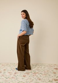 Blauer, kurzgeschnittener, strukturierter Pullover mit kurzen Ärmeln, kombiniert mit braunen Cord-Hosen mit weitem Bein. Vor einem floralen Muster auf dem Boden.