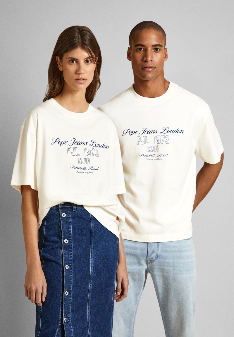 Zwei Modelle tragen übergroße cremefarbene T-Shirts mit navyfarbenen Grafiken. Die Frau kombiniert ihres mit einem blauen Jeansrock, der weiße Knöpfe hat; der Mann trägt helle Jeans.