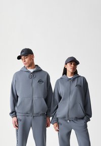 Completo felpa grigia con zip, cappuccio e dettaglio logo. Il tessuto appare morbido con una vestibilità comoda. Inclusi cappellini coordinati.