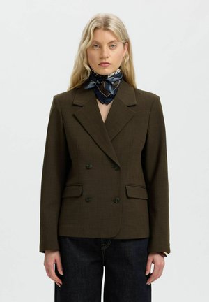Femme blonde portant un blazer marron foncé croisé, un foulard à motifs bleu marine autour du cou et un jean foncé, faisant face à la caméra sur fond blanc.