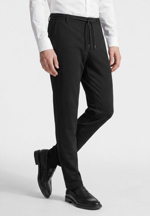 Man draagt zwarte slimfit broek met aantrekkoord in de taille, zwarte leren loafers en een wit overhemd met lange mouwen, staand met één been over het andere geslagen.