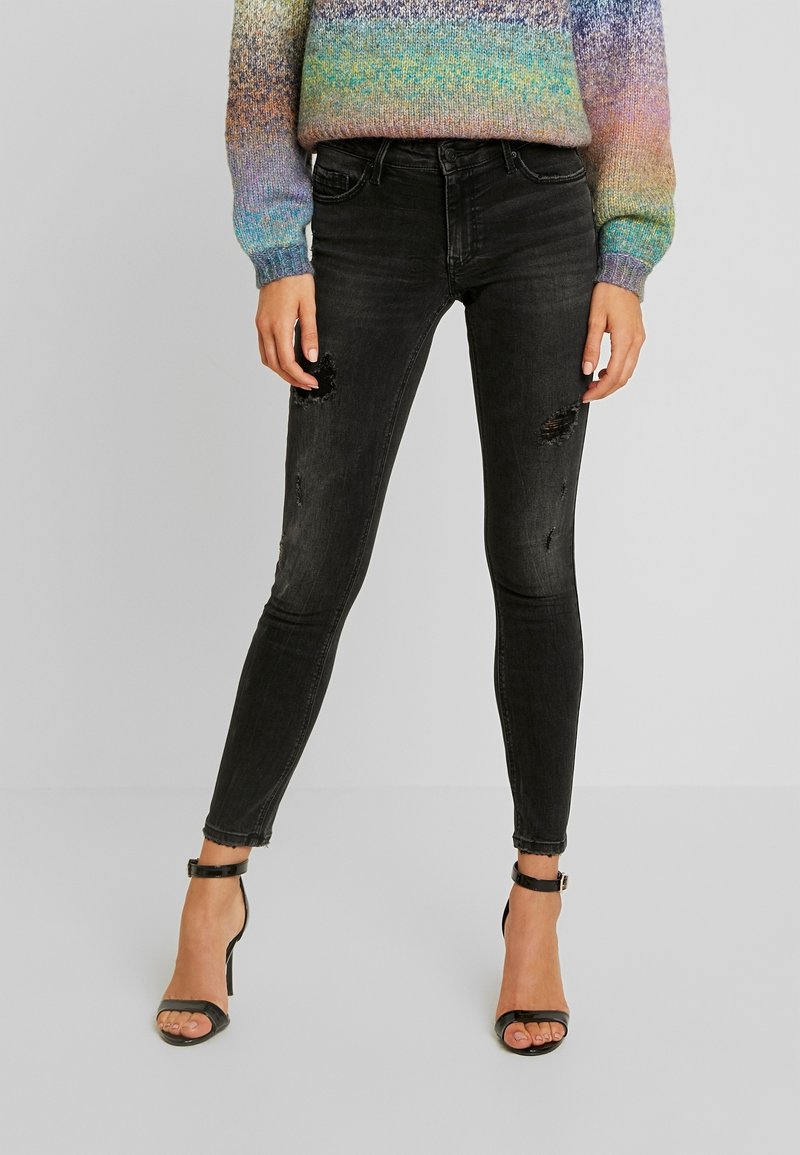 Femme portant un jean skinny noir déchiré, un pull en tricot multicolore et des sandales noires à talons hauts avec brides à la cheville, debout devant un fond uni.