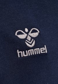 Marineblauem Stoff mit gesticktem Logo, das ein abstraktes Bienenmotiv zeigt. "hummel" ist in Hellgrau unter dem Logo gestickt.