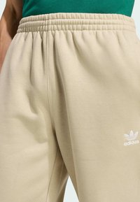 Beżowe spodnie dresowe z gromadzącym się elastycznym pasem. Wykonane z miękkiego materiału, z małym białym logo Adidas na dolnej części nogawki.