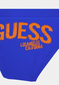 Kék úszónadrág sima anyagból. Az elülső részen narancssárga "GUESS" logó és "LOS ANGELES CALIFORNIA" felirat található, egy kopott stílusú betűtípussal.