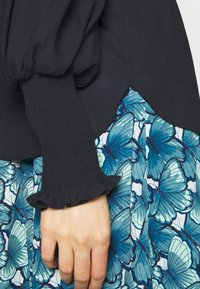 Main reposant à côté d'une jupe plissée avec imprimé papillon bleu, portant un haut noir à manchette texturée et volantée.