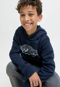 Lächelnder Junge mit lockigem Haar, der einen dunkelblauen Hoodie und Jeans trägt, sitzt mit einem Arm auf seinem Knie vor einem schlichten weißen Hintergrund.
