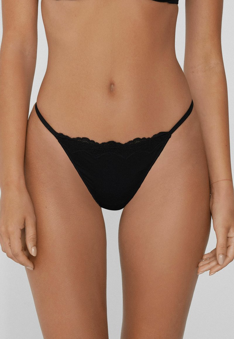 Femme portant un string noir à bord en dentelle, découpée de la poitrine aux cuisses supérieures, se tenant devant un fond clair et uni.