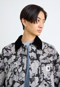 Giovane con capelli neri corti girato di lato, indossando una giacca camouflage nero-grigia con colletto nero e una camicia azzurra chiara sotto.