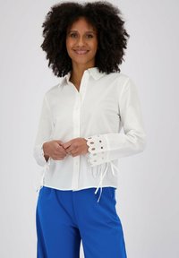 Camicia bianca con bottoni e maniche lunghe, caratterizzata da polsini a scallop e dettagli in occhio di pernice. Abbinata a pantaloni blu in morbido cotone.