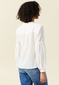 Blusa bianca, leggera, con maniche lunghe e ampie e una trama sottile. Il retro presenta un design a spallina e un orlo dritto. Abbinata a jeans blu.